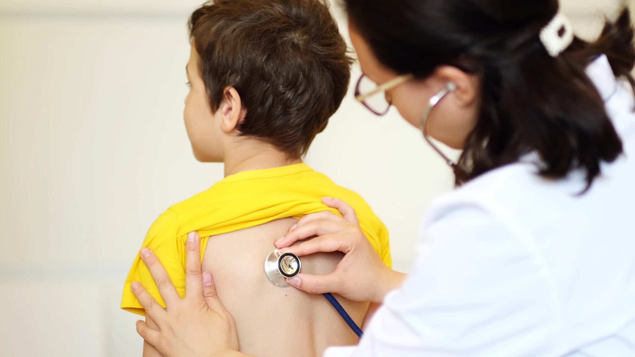 A female healthcare professional holding a stethoscope to a male child's back to listen to their lungs.